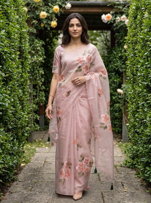 A woman stands on a stone path between tall green hedges wearing a light pink floral saree with a matching blouse. The saree has pink and peach flowers with green leaves and dark green tassel ties.
