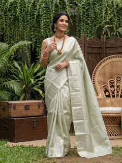 Woman in a light green saree standing outdoors with plants and a wooden chair in the background