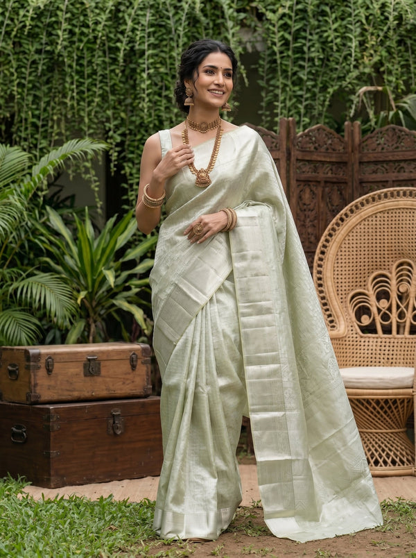 Woman in a light green saree standing outdoors with plants and a wooden chair in the background
