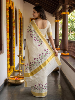 A woman wearing a white saree with purple floral embroidery and gold borders, standing on a traditional porch decorated with marigold garlands.