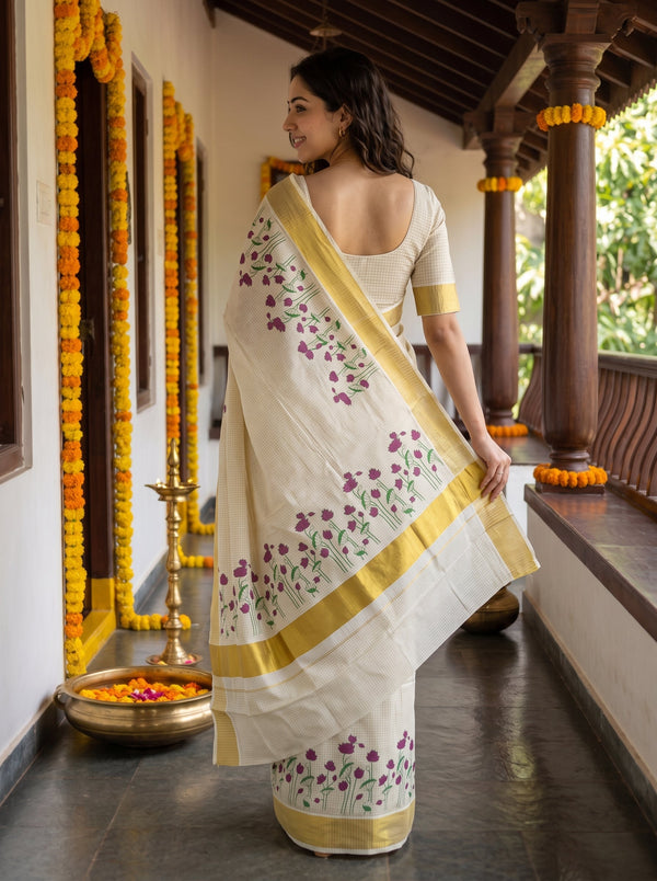 A woman wearing a white saree with purple floral embroidery and gold borders, standing on a traditional porch decorated with marigold garlands.