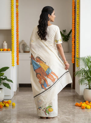A woman wearing a cream-colored cotton saree with a colorful illustration of children and a lemon on the border, paired with a matching cream kurta, standing in a decorated indoor space with marigold garlands.