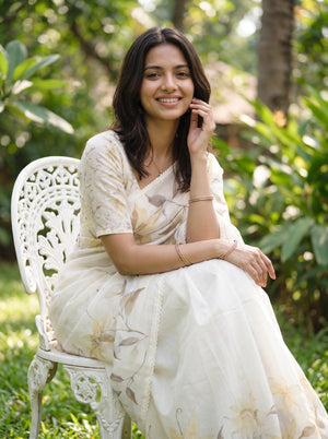 A woman wearing a cream-colored floral printed saree sits on a white garden chair outdoors, surrounded by green foliage.