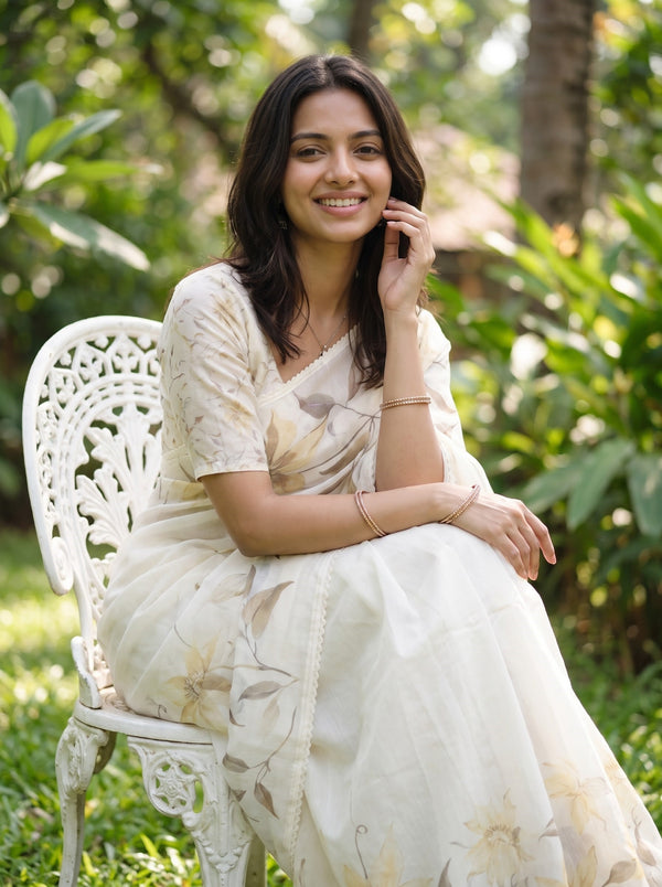 A woman wearing a cream-colored floral printed saree sits on a white garden chair outdoors, surrounded by green foliage.