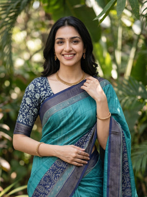 A woman wearing a teal and navy blue saree with floral patterns on the sleeves, standing outdoors against a lush green background.