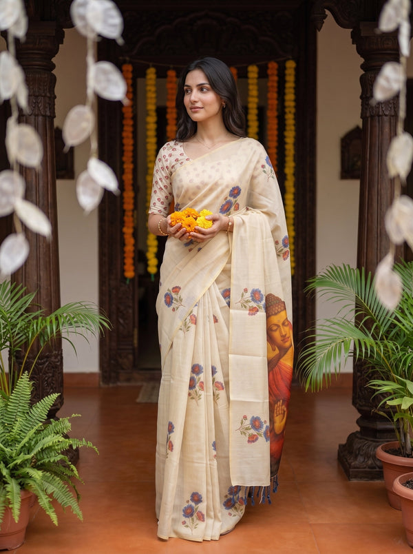 A woman wearing a cream-colored printed saree with floral motifs and a Buddha print on the pallu, standing in a decorated courtyard and holding yellow marigold flowers.