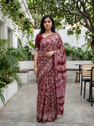 A woman wearing a maroon and beige patterned saree with a matching maroon blouse, standing in an outdoor courtyard with plants and tables in the background.
