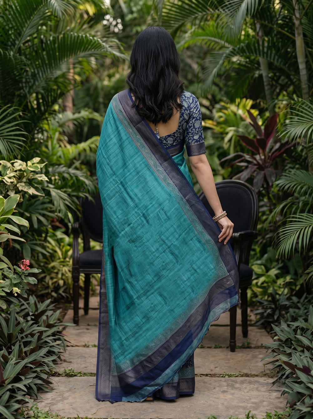 A woman stands outdoors in a lush tropical garden wearing a teal and navy blue patterned sari. The sari has a solid teal body with a dark navy border and pallu featuring intricate floral motifs, paired with a matching navy blue blouse.