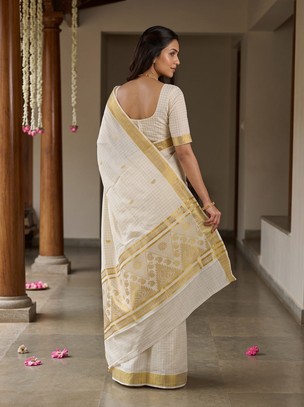 A woman wearing a cream-colored cotton saree with a gold border and matching cream blouse, standing in a traditional corridor with wooden pillars.