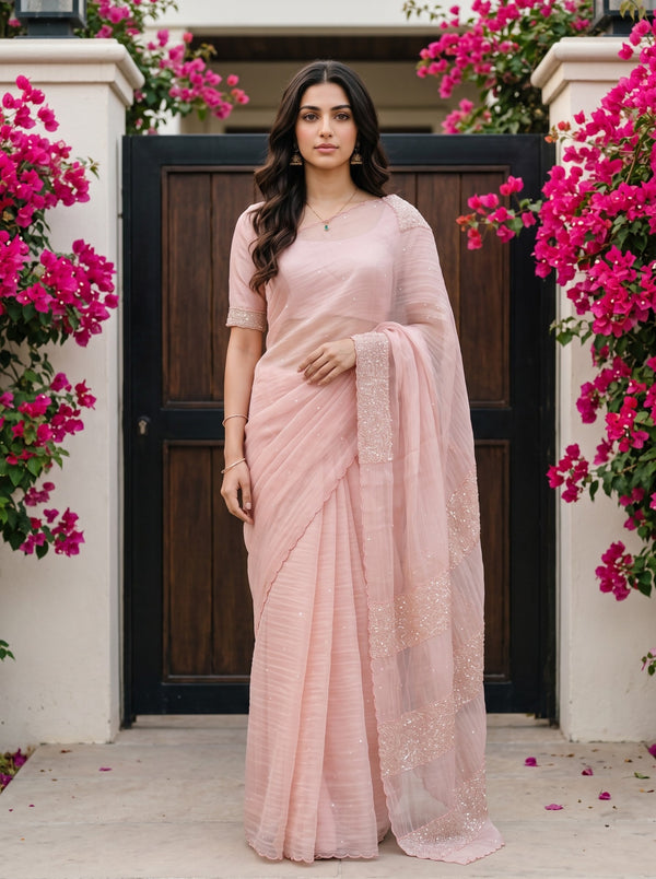 A woman wearing a light pink traditional sari with matching blouse stands in front of a dark wooden door framed by white pillars and abundant bright pink flowers.