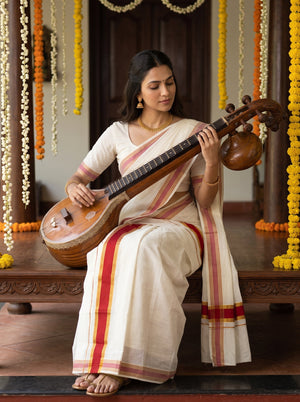 A woman seated on a wooden bench holding a traditional stringed instrument, wearing a cream-colored saree with red and gold stripes. The background features hanging marigold garlands and a dark wooden interior.