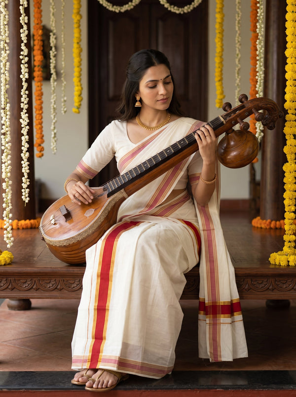 A woman seated on a wooden bench holding a traditional stringed instrument, wearing a cream-colored saree with red and gold stripes. The background features hanging marigold garlands and a dark wooden interior.