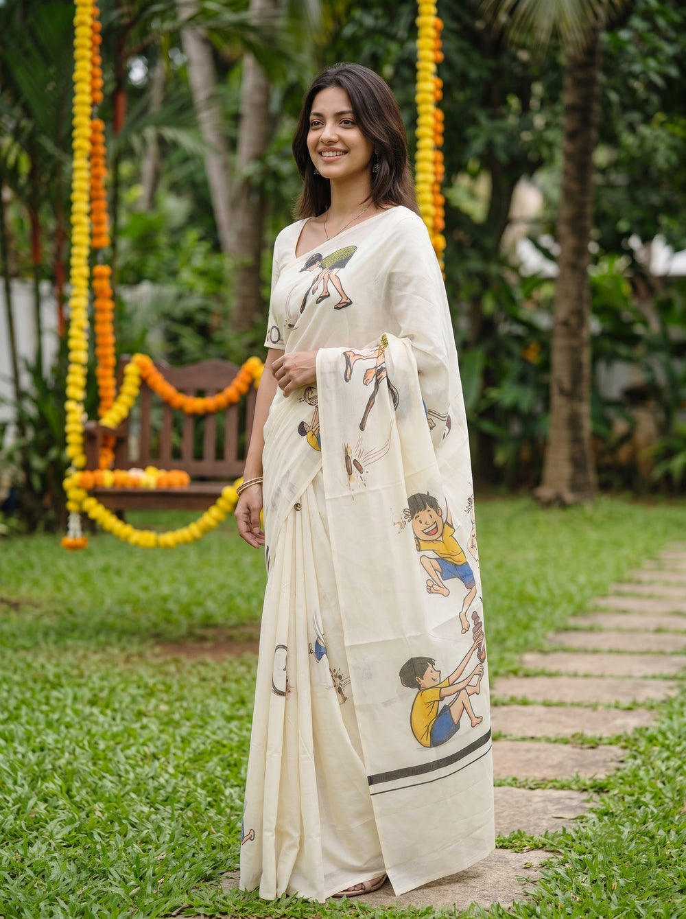 A woman viewed from behind wearing a cream-colored cotton saree with colorful traditional Indian folk art illustrations, standing on a stone path in a garden.