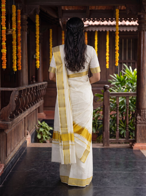 A woman viewed from behind wearing a white saree with gold borders, standing on a dark tiled porch in front of a traditional wooden building with yellow marigold garlands hanging from the ceiling.