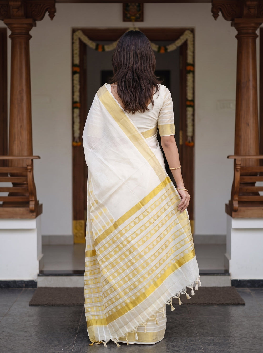 A woman modeling a cream-colored cotton saree with yellow stripes and a matching yellow border, standing in front of a traditional doorway with wooden pillars.