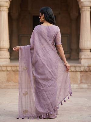 A woman wearing a lavender purple saree with a matching blouse, shown from behind in front of a stone architectural background with pillars.
