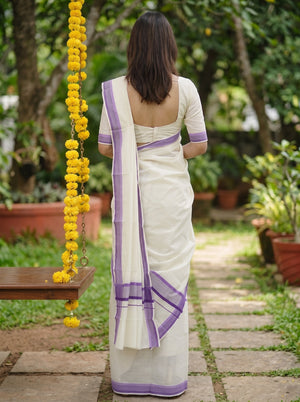A woman viewed from behind wearing a white saree with purple border accents, standing on a stone path in a garden with a swing and yellow flower garlands nearby.