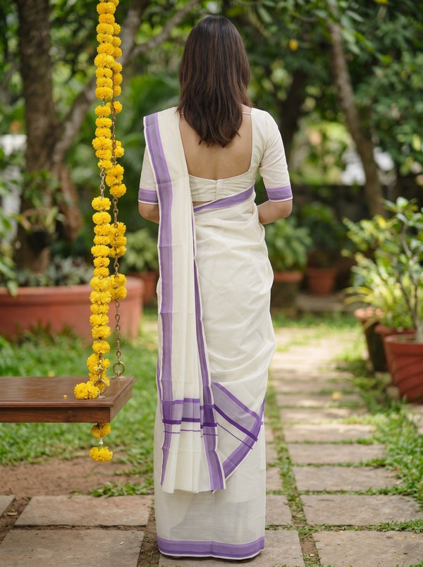 A woman viewed from behind wearing a white saree with purple border accents, standing on a stone path in a garden with a swing and yellow flower garlands nearby.