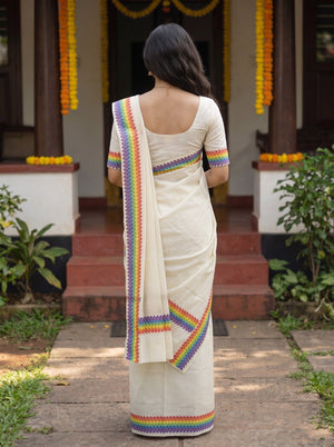 A woman viewed from behind wearing a cream-colored saree with colorful rainbow-patterned borders, standing in front of a decorated doorway with steps and plants.