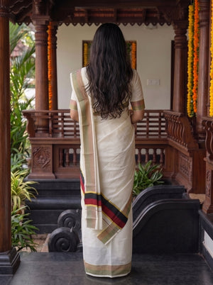 A woman viewed from behind wearing a cream-colored cotton saree with a striped border in red, green, and gold, standing in front of a traditional wooden pavilion.