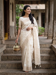A woman wearing a cream-colored cotton saree with a matching printed blouse stands on stone steps in a traditional courtyard, holding a small basket of flowers.