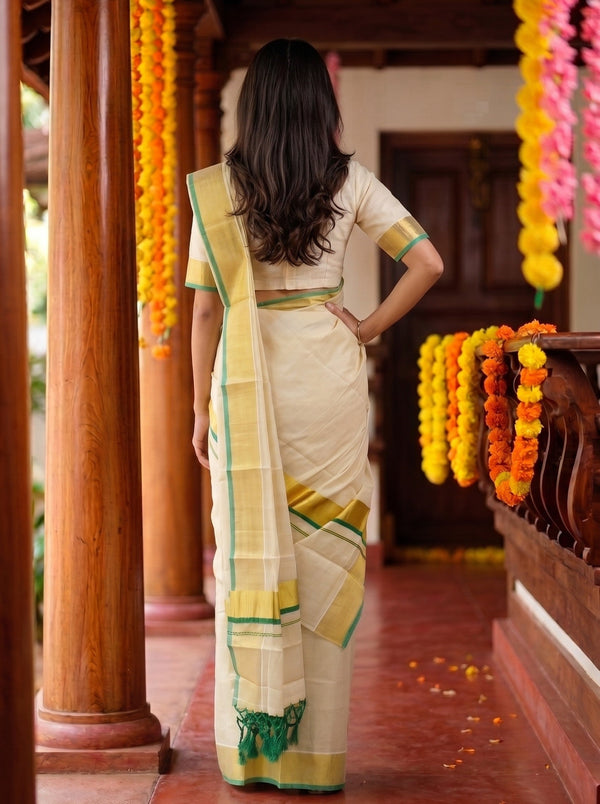 A woman viewed from behind wearing a cream-colored traditional saree with yellow and green borders, standing on a wooden porch decorated with yellow and orange flower garlands.