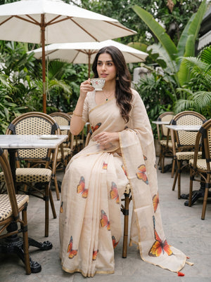 A woman seated at an outdoor cafe table wearing a light beige saree with colorful butterfly prints, holding a teacup. The background features green foliage, patio umbrellas, and woven chairs.