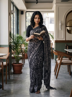 A woman wearing a black and gray patterned sari stands in a cafe reading a book. The sari features a black base with intricate gray and white floral motifs, paired with a solid black blouse.