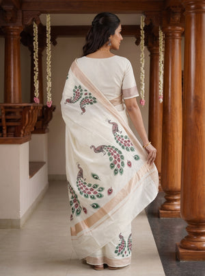 A woman wearing a white saree with peacock and floral embroidery, standing in a corridor with wooden pillars.