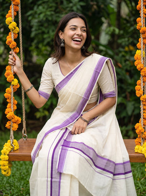 A woman wearing a cream-colored cotton saree with purple borders sits on a wooden swing decorated with orange marigold garlands, set against a lush green garden background.