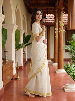 A woman wearing a white cotton saree with gold border accents, standing in a traditional courtyard with wooden pillars and tiled floors.