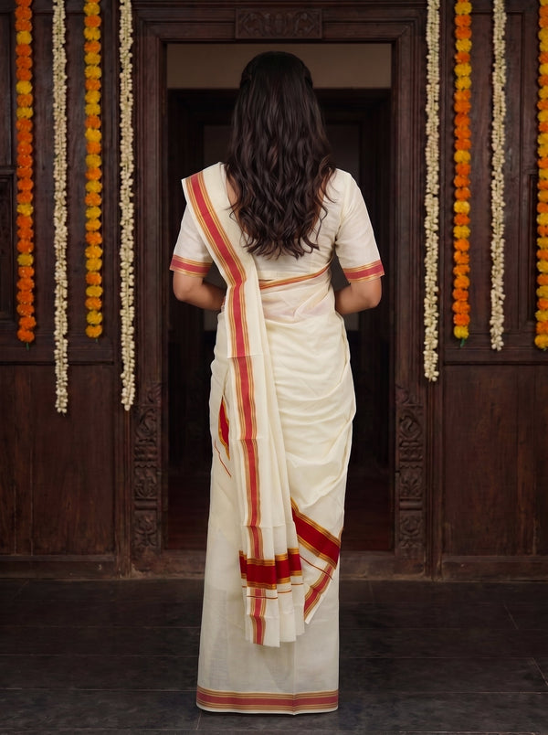 A woman viewed from behind wearing a cream-colored cotton saree with red and orange stripes, standing in front of a dark wooden doorway decorated with hanging marigold garlands.