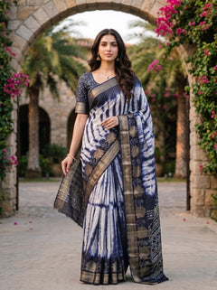 A woman models a blue and white tie-dye saree with a metallic gold border, standing in front of a stone archway decorated with greenery and pink flowers.