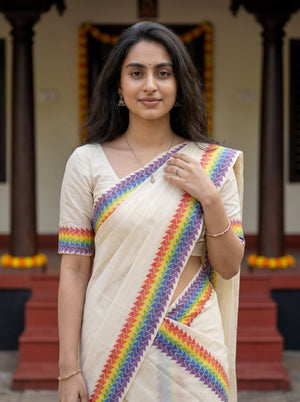 A woman wearing a cream-colored sari with vibrant rainbow patterned borders, standing in front of a traditional building with dark pillars.