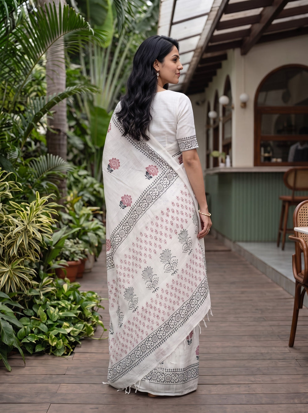 A woman modeling a white cotton saree with pink floral prints and black geometric borders, standing on a wooden deck in a tropical garden setting.