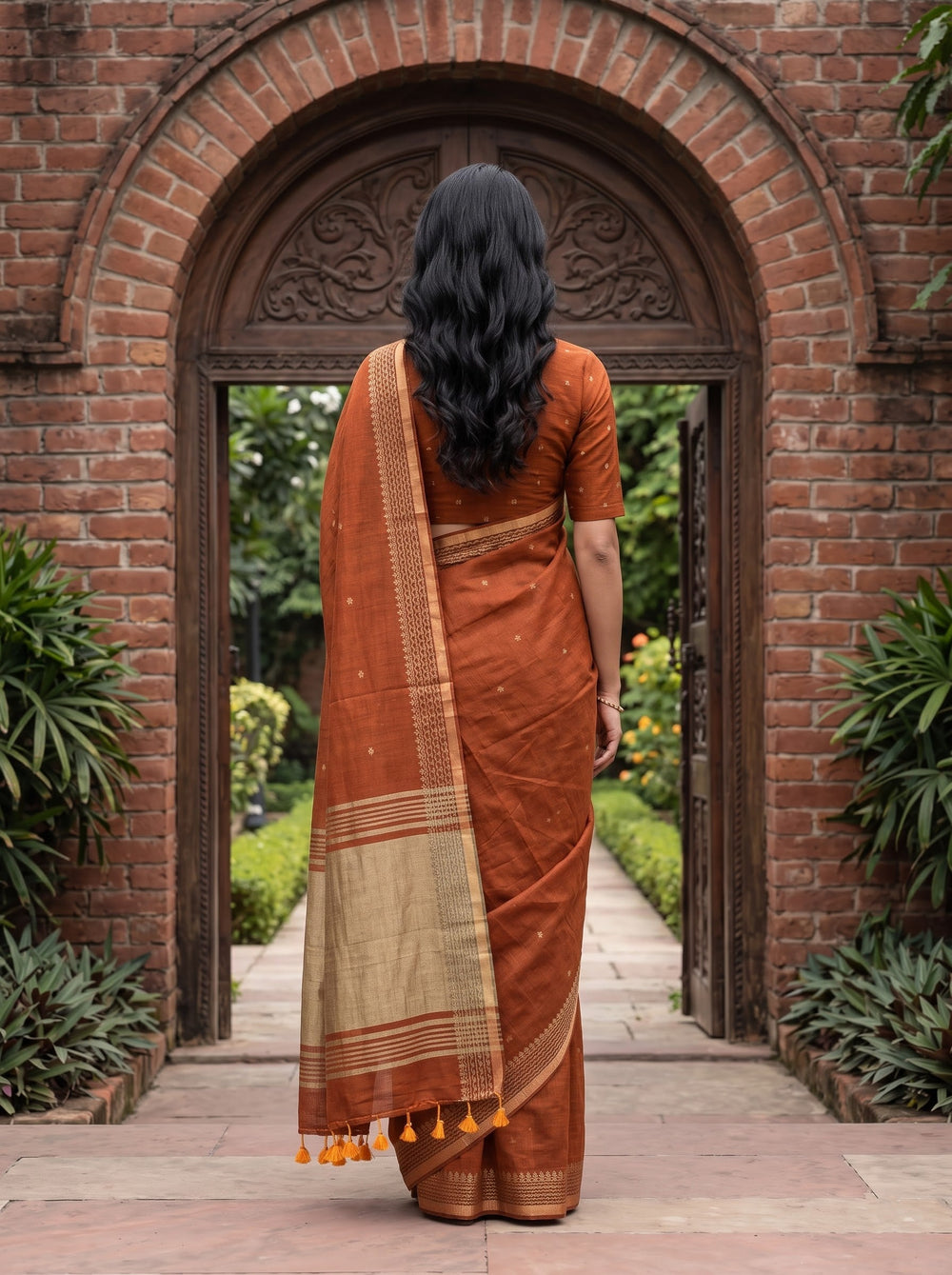 A woman wearing a rust-orange and beige saree stands centered in front of a brick archway with greenery on both sides. The saree has a warm rust-orange body with a beige border and small metallic-looking motifs, and the blouse is a matching rust-orange color.