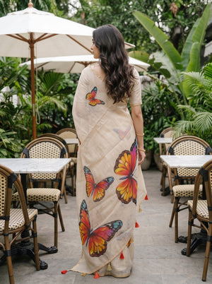 A woman stands in a garden cafe wearing a beige sari decorated with large colorful butterfly prints in orange, yellow, and blue, with small red tassel accents.