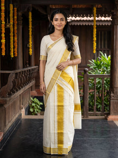 A woman wearing a traditional Indian saree stands on a wooden porch. The saree is off-white/cream with a yellow-gold border and pallu, paired with a matching cream blouse with yellow-gold trim.