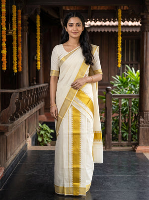 A woman wearing a traditional Indian saree stands on a wooden porch. The saree is off-white/cream with a yellow-gold border and pallu, paired with a matching cream blouse with yellow-gold trim.