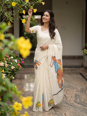 A woman wearing a cream-colored saree with colorful printed illustrations stands in a garden courtyard with yellow flowers in the foreground.