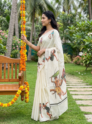 A woman wearing a cream-colored saree with traditional Indian folk art prints stands in a lush green garden next to a wooden swing decorated with orange and yellow marigold garlands.