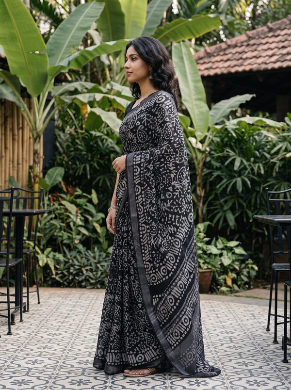 A woman wearing a black and white patterned saree stands in profile in an outdoor garden patio setting with tropical plants and a tiled floor.