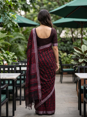 A woman viewed from behind wearing a dark red and black patterned saree with a deep purple border and a solid dark purple blouse, standing in an outdoor patio setting with green umbrellas and plants.