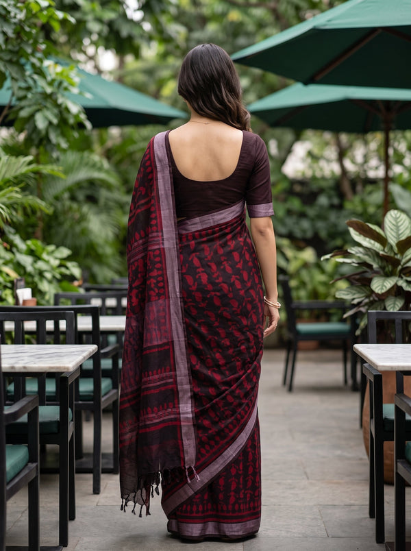 A woman viewed from behind wearing a dark red and black patterned saree with a deep purple border and a solid dark purple blouse, standing in an outdoor patio setting with green umbrellas and plants.