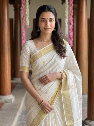 A woman wearing a cream-colored saree with a subtle grid pattern and a golden-yellow border, standing in a corridor with wooden pillars.