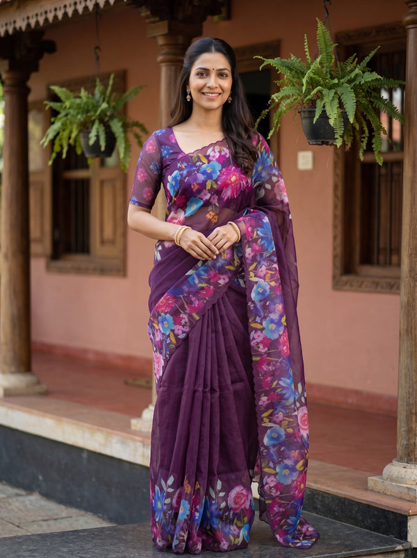 A woman wearing a purple floral printed saree stands on the steps of a traditional building with pink walls and hanging green plants.