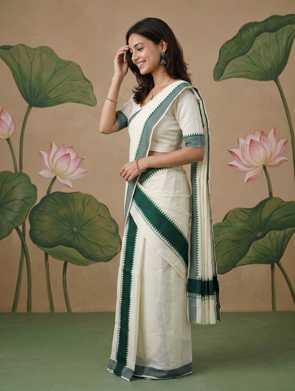 A woman modeling a cream-colored saree with dark green borders and a matching green blouse, standing in front of a beige backdrop decorated with painted lotus leaves and flowers.