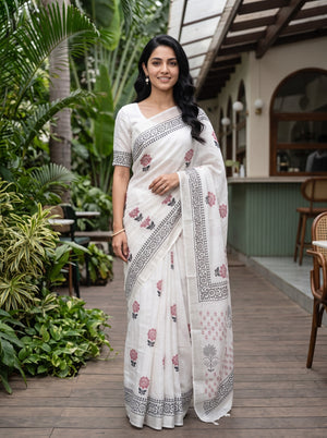 A woman modeling a white cotton saree with pink floral prints and black geometric borders, standing on a wooden deck in a tropical garden setting.