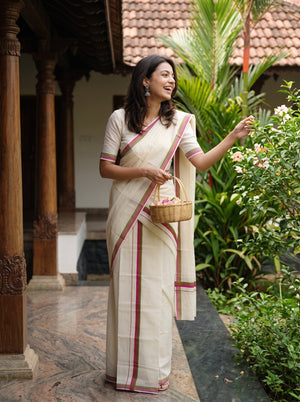 A woman wearing a cream-colored cotton saree with red stripes and a matching short-sleeve blouse stands on a tiled porch, holding a small woven basket next to lush green plants and flowers.