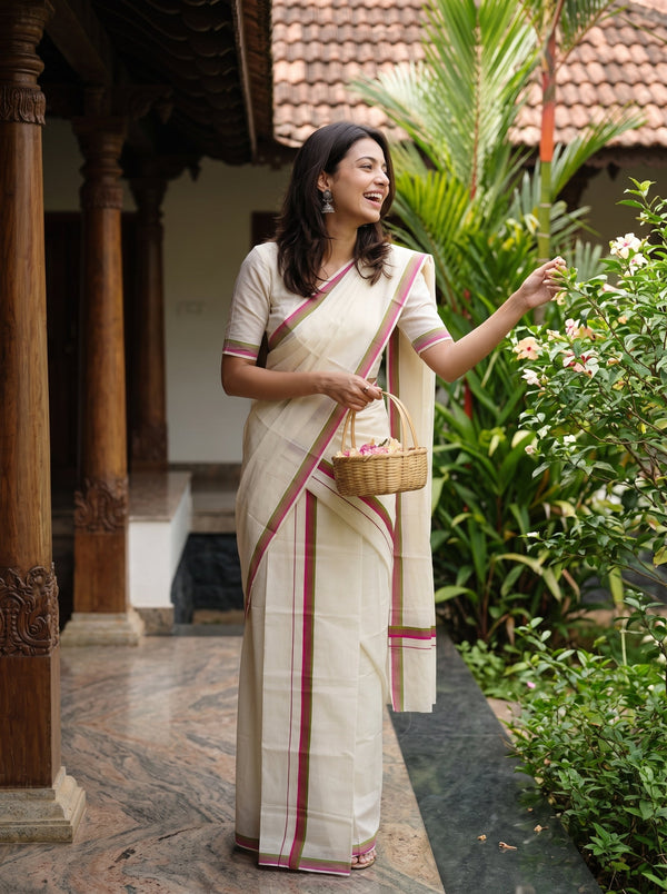 A woman wearing a cream-colored cotton saree with red stripes and a matching short-sleeve blouse stands on a tiled porch, holding a small woven basket next to lush green plants and flowers.